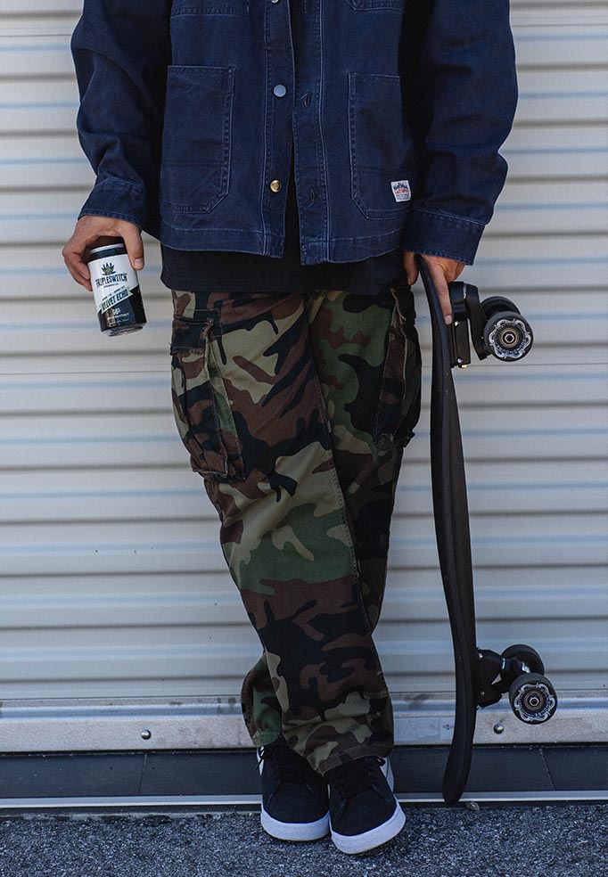 Person wearing a blue jacket and camouflage pants holding a bottle of echo velvet beer and skateboard against a metallic background