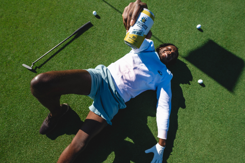 Man lying on golf green holding a can of Napleswitch Uno Mas THC beverage near golf balls, putter, and flag